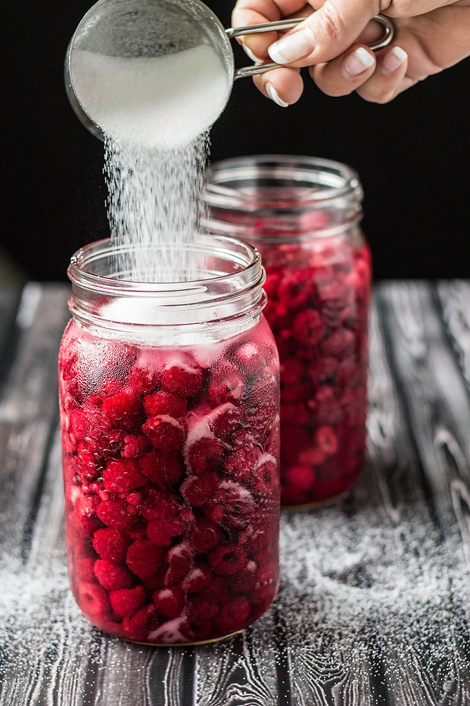 pouring sugar into mason jars full of raspberries and vodka to make raspberry liqueur