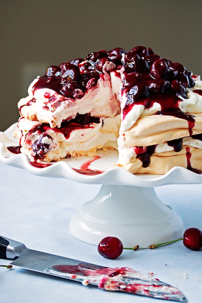 layered pavlova recipe with whipped cream in the middle and cherry compote on top. a slice is missing from the dessert