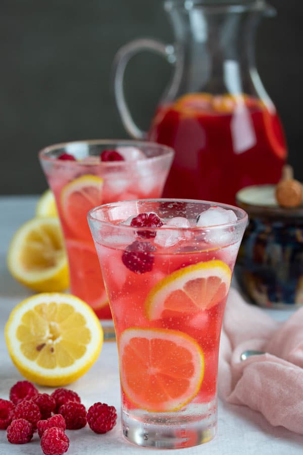 a glass of hard raspberry lemonade with another cocktail and pitcher in the background