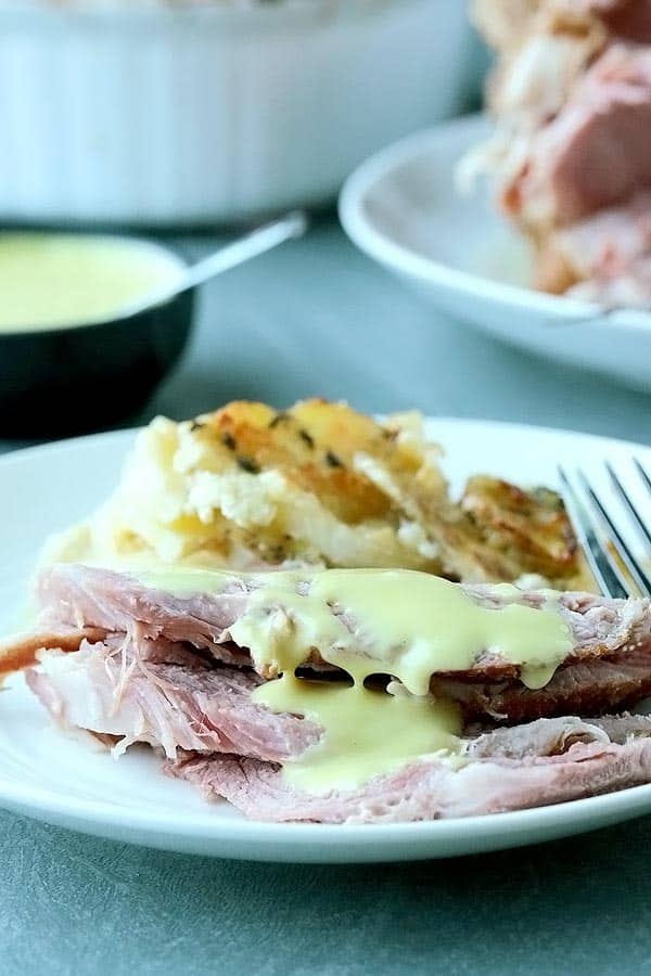 a plate of ham with mustard sauce drizzled on top and scalloped potatoes in the back